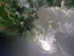 Image of the Deepwater Horizon oil spill in the Gulf of Mexico as shot by NASA from space, May 24, 2010. The images shows the peninsula of southern Louisiana where New Orleans is located, with the channels of the Mississippi River clearly visible flowing into the Gulf from the land in the upper left. Slighly lower from center there is a large white swirly object that looks a bit like a cloud but trails off to the east-northeast; this is spilled oil covering the surface of the ocean over hundreds of square miles.