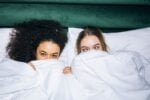 Two young girls lying on a white bed with a green velour blanket or long pillow across the top of the bed and photo.  They have a white blanket pulled up over them so only their eyes, a bit of their hands, and their foreheads and hair are visible; the girl on the left has full black or dark brown hair that is very curly, while the girl on the right has blonde straight hair and her eyes are crossed.  Both girls appear to be smiling, and the overall aesthetic of the photo is youth, fun, and female with just the barest hint of "naughty."